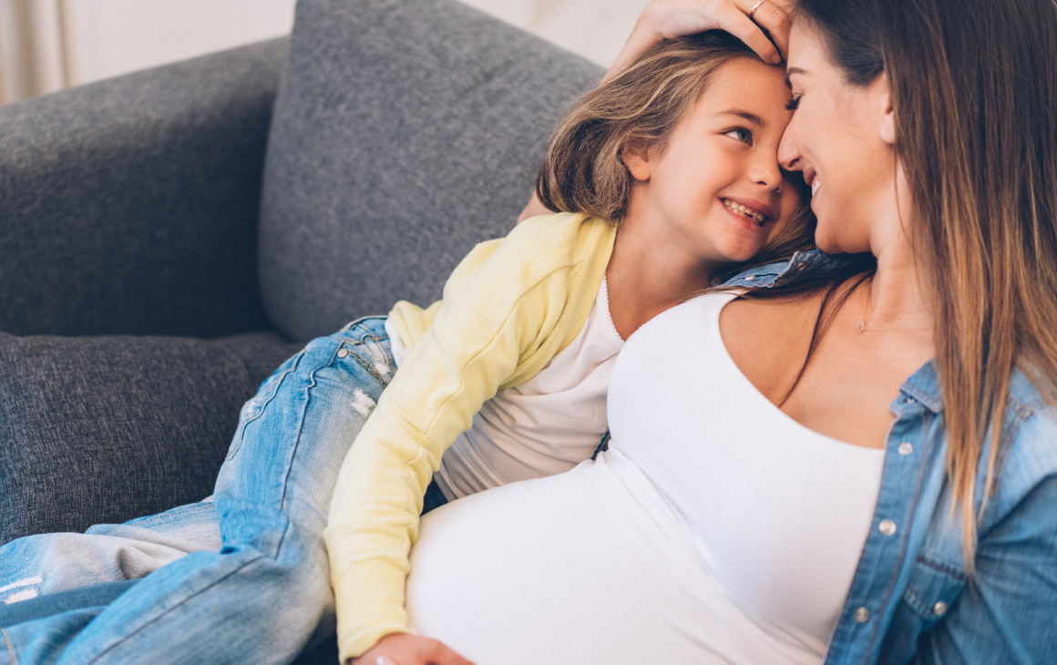 Mãe grávida a sorrir carinhosamente com a filha no sofá.